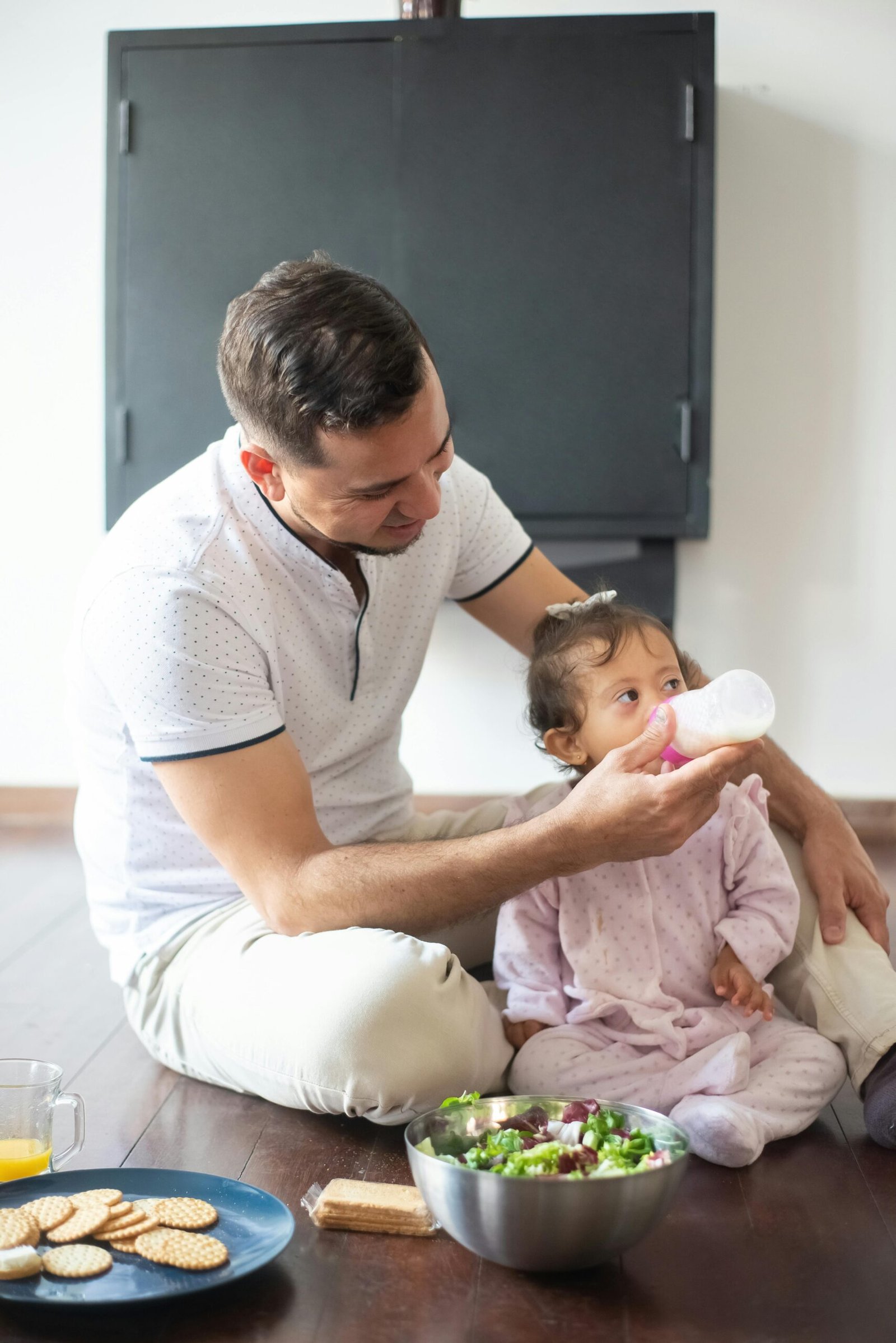 A father lovingly feeds his baby daughter while sitting together at home indoors.